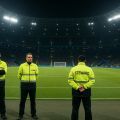 Personal del dispositivo de seguridad y emergencias en un estadio lleno durante un partido nocturno, con chalecos reflectantes y presencia estratégica en el perímetro del campo.